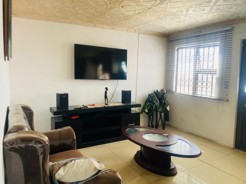 A living room featuring tiled flooring, decorative ceiling tiles, and a window fitted with security bars and venetian blinds.