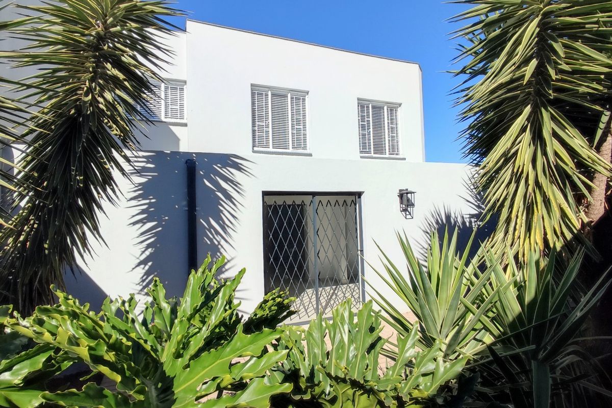 Rear elevation of the townhouse featuring white rendered walls, window shutters, and a secure metal gate entrance framed by established garden foliage.