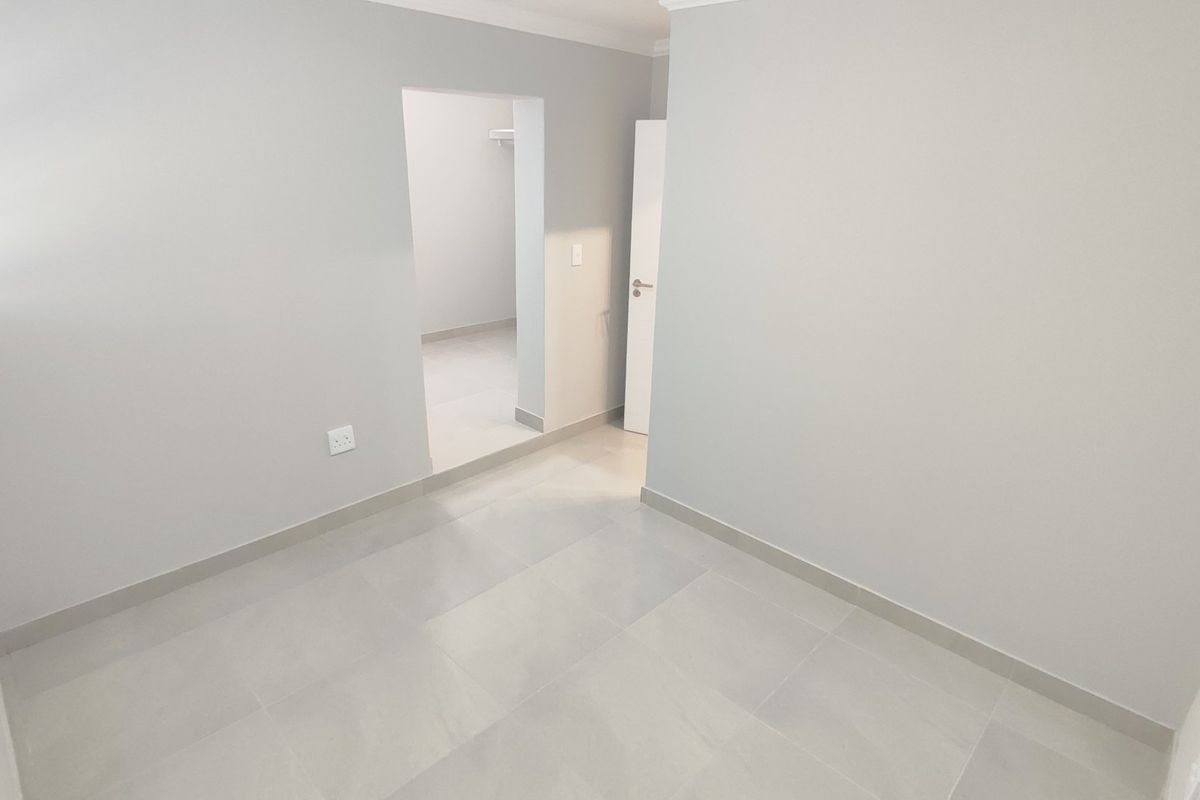 A bright, neutral, and empty bedroom featuring modern tiled flooring, clean finishes, and an adjoining walk-in closet or storage area.