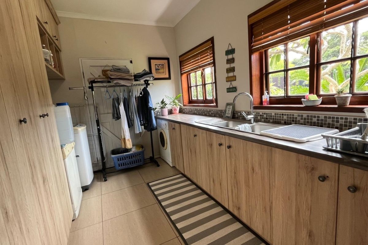 Functional laundry room featuring wood-look cabinetry, double basin sink, mosaic tile backsplash, and tiled flooring with ample natural light from timber-framed windows.