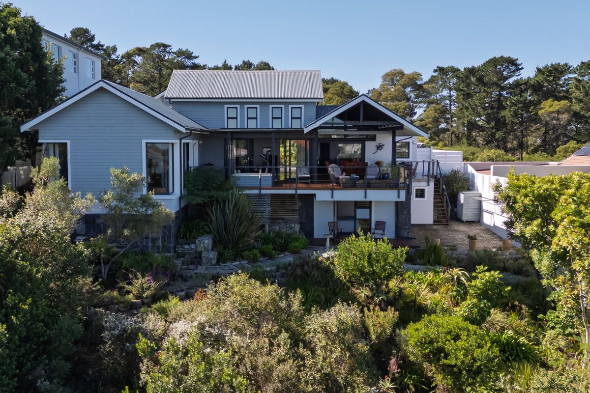 Rear view of a multi-level house featuring timber decking, weatherboard cladding, a corrugated metal roof, and an established landscaped garden.