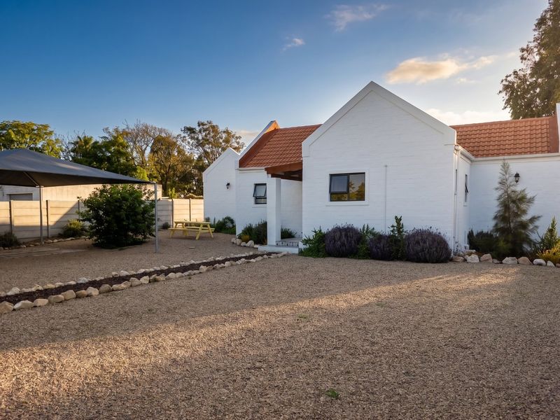 A well-maintained single-story home featuring a white painted brick facade, terracotta roof tiles, and a spacious gravel driveway with a shaded parking area.