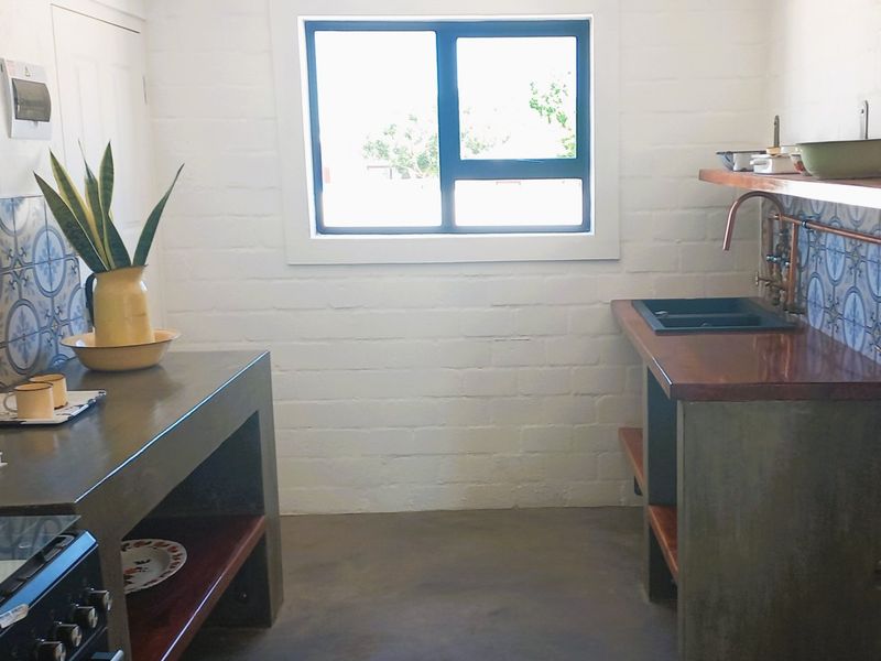 A rustic-style kitchen featuring concrete countertops, exposed copper plumbing, decorative tile backsplashes, and a built-in stove with timber shelving.