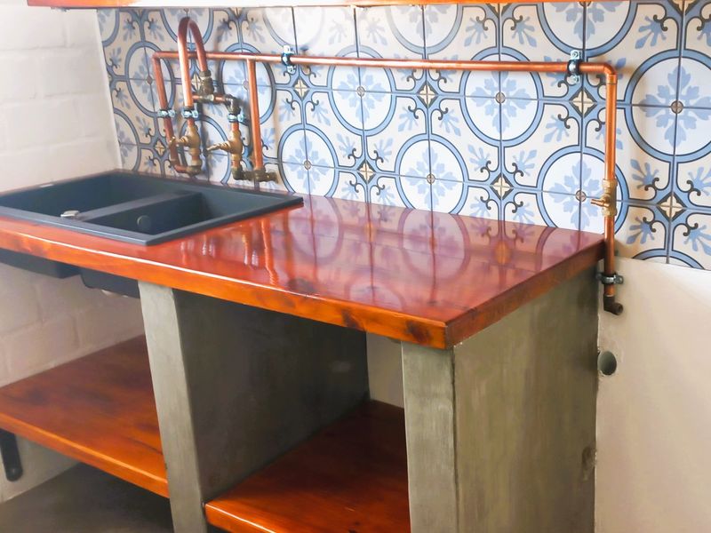 A modern kitchen prep area featuring a concrete base, timber countertop, undermount sink, and decorative patterned wall tiles with exposed copper plumbing.