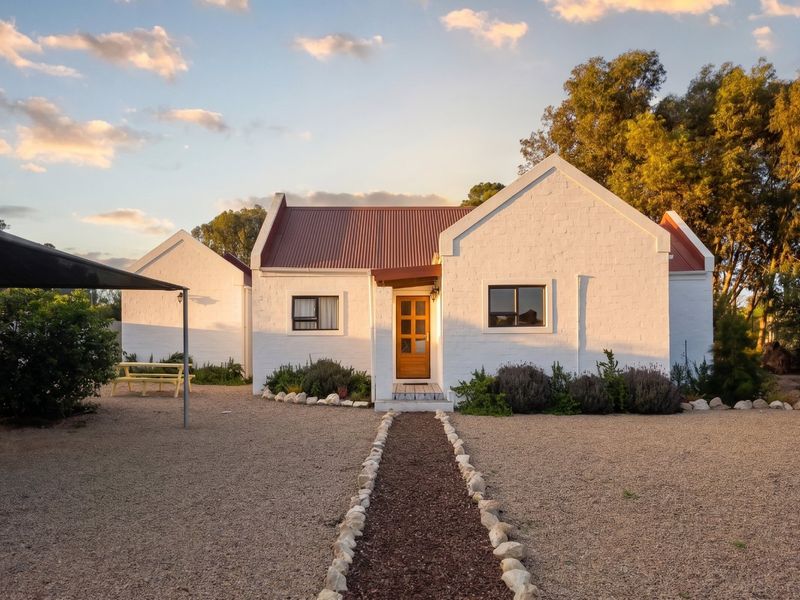 Charming single-story cottage featuring a white painted brick facade, corrugated metal roof, and a gravel driveway with a landscaped stone-bordered path.