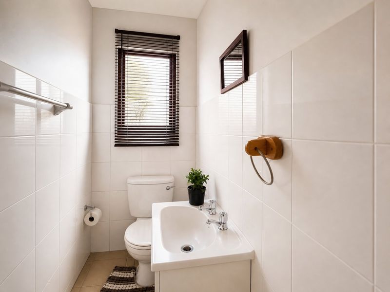 A clean and functional guest bathroom featuring white wall tiles, a pedestal washbasin, and a window with a venetian blind.