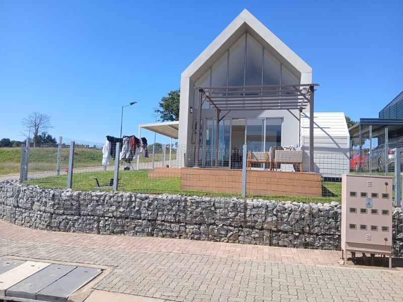 Modern gabled house exterior featuring a gabion retaining wall, timber pergola, and large glass sliding doors opening to a small lawn area.