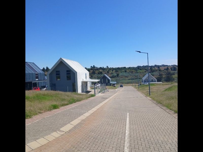 A view of the paved street approach within a residential estate, featuring modern homes with metal roofing and surrounding open landscape.