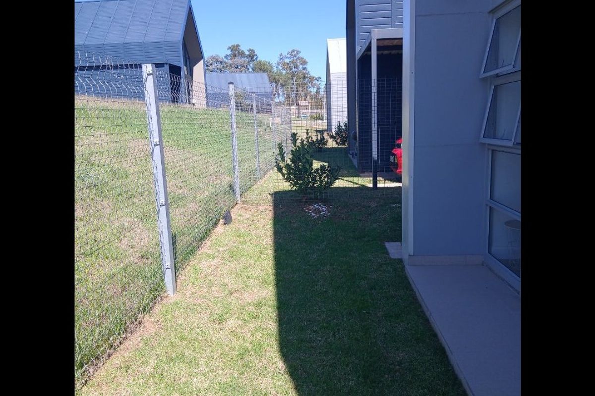 A side yard view showing a narrow lawn area, wire mesh fencing, and the side elevation of the modern residential building.