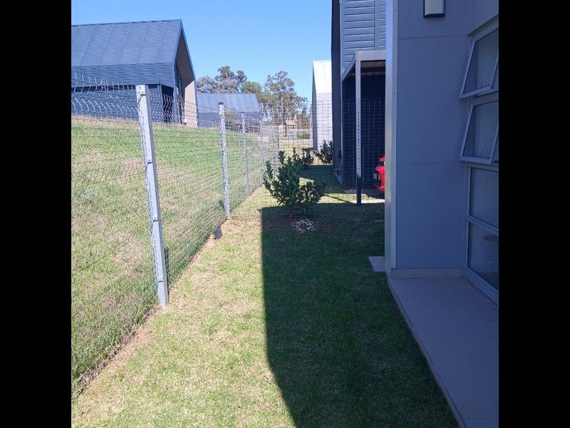 A side yard view showing a narrow lawn area, wire mesh fencing, and the side elevation of the modern residential building.
