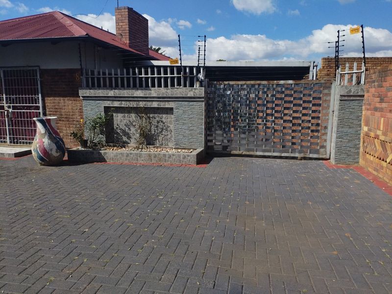 Front exterior view featuring a paved driveway, secure metal gate, electric fencing, and decorative stone cladding on the boundary wall.