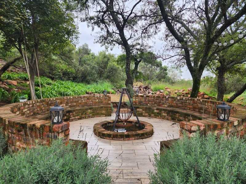 A tranquil outdoor garden area featuring a circular brick fire pit, curved built-in brick seating, and paved flooring surrounded by mature trees.