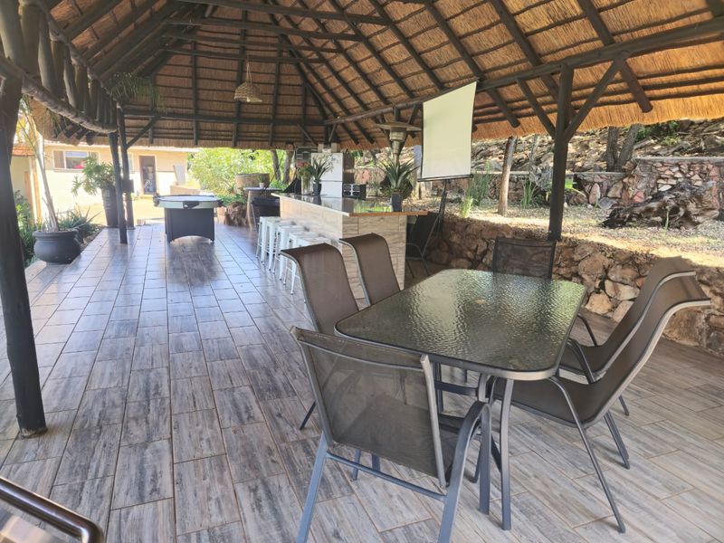 Spacious covered outdoor entertainment area featuring a thatched roof, timber support beams, tiled flooring, and a built-in bar counter set against a stone retaining wall.