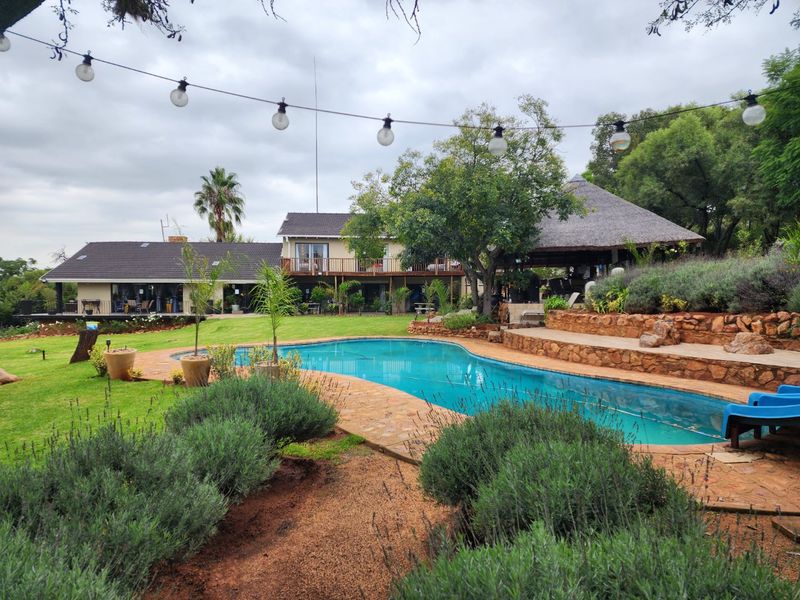 Spacious outdoor pool area featuring a swimming pool, stone terracing, a thatched lapa, and a large garden with a view of the main building.