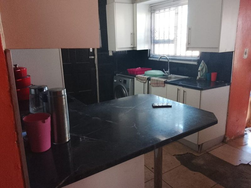 A compact kitchen featuring dark laminate countertops, white cabinetry, and a tiled splashback, requiring some cosmetic updates to the flooring and wall finishes.