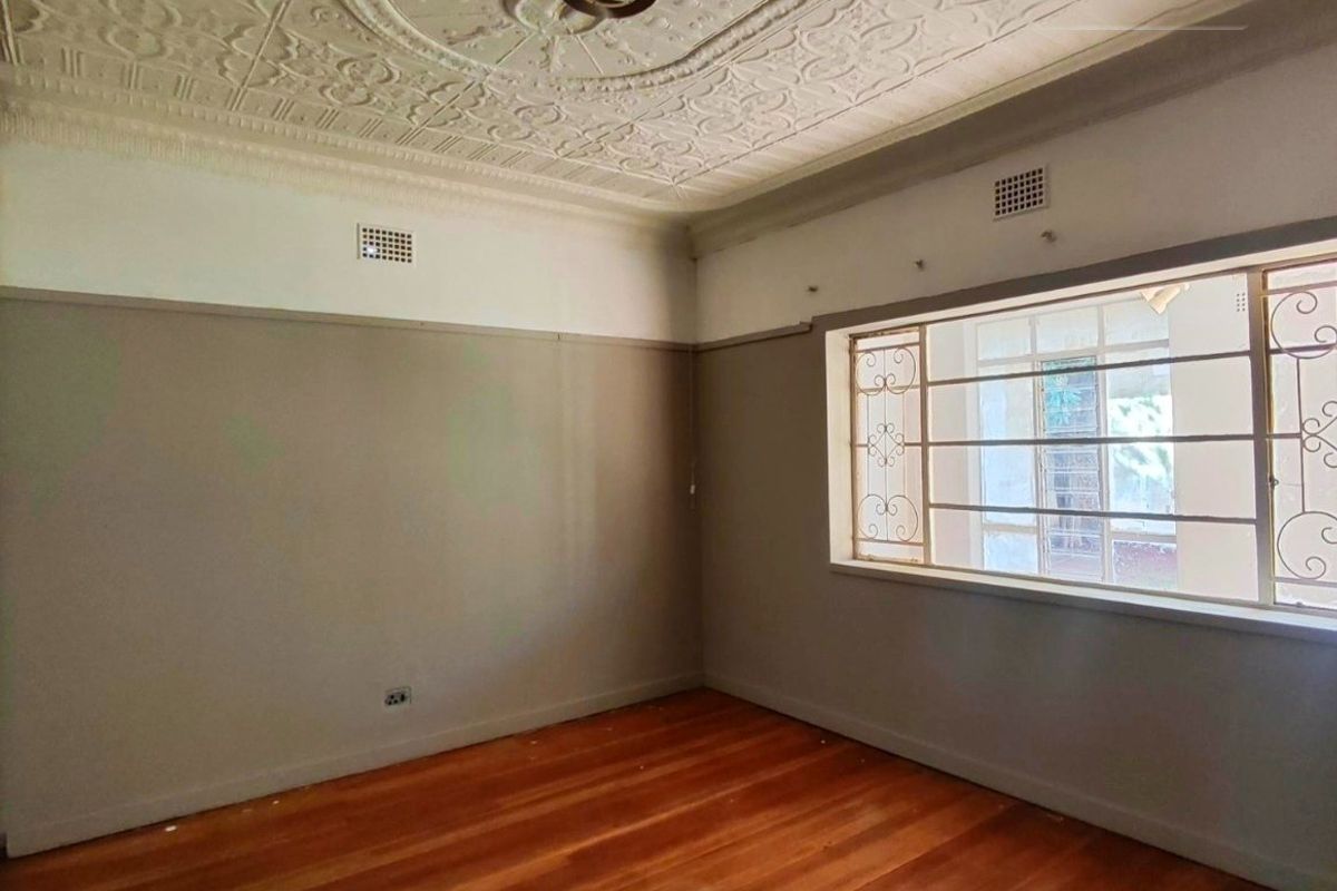 An empty bedroom featuring original pressed metal ceilings, hardwood flooring, and security bars on the window, offering a classic architectural foundation.