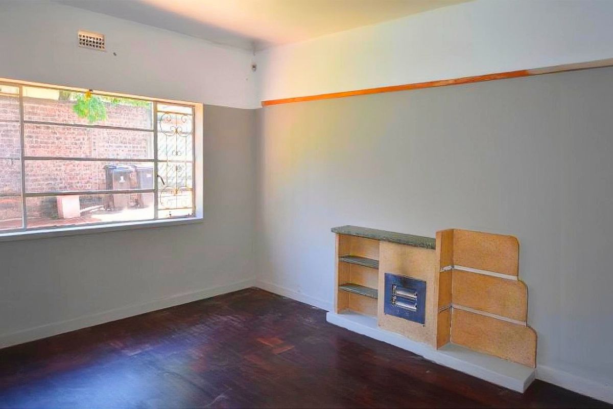 An empty living room featuring original hardwood flooring, a steel-framed window, and a built-in shelving unit with a central fireplace insert.