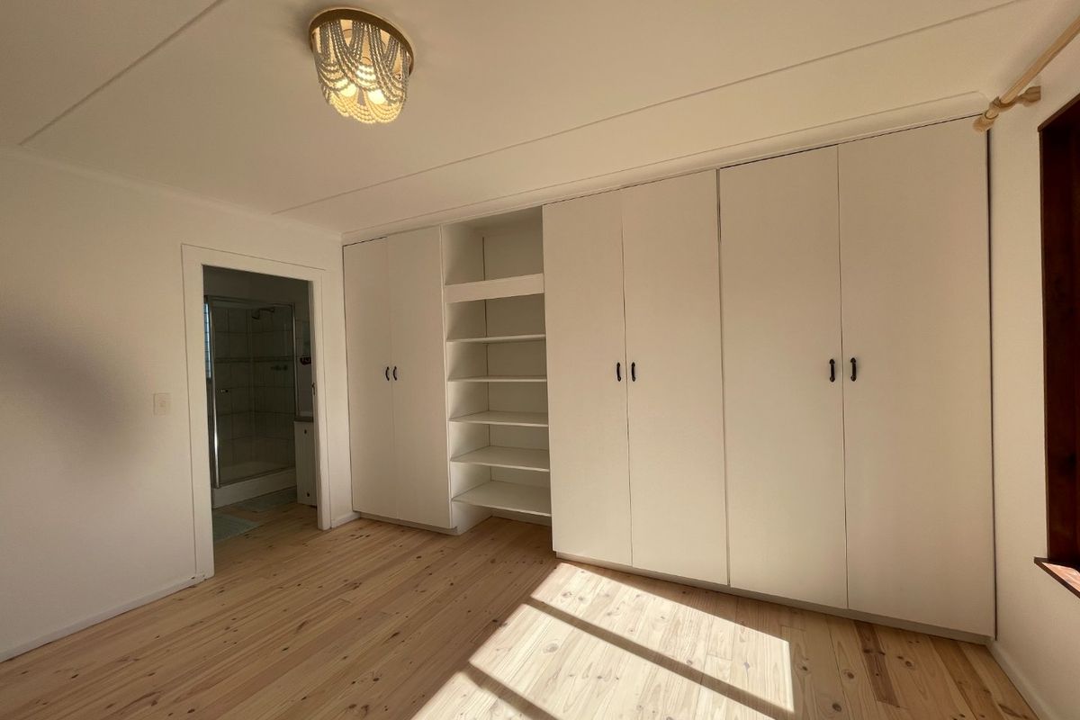 A bright bedroom featuring polished timber flooring, extensive built-in storage cupboards with open shelving, and direct access to an en-suite bathroom.