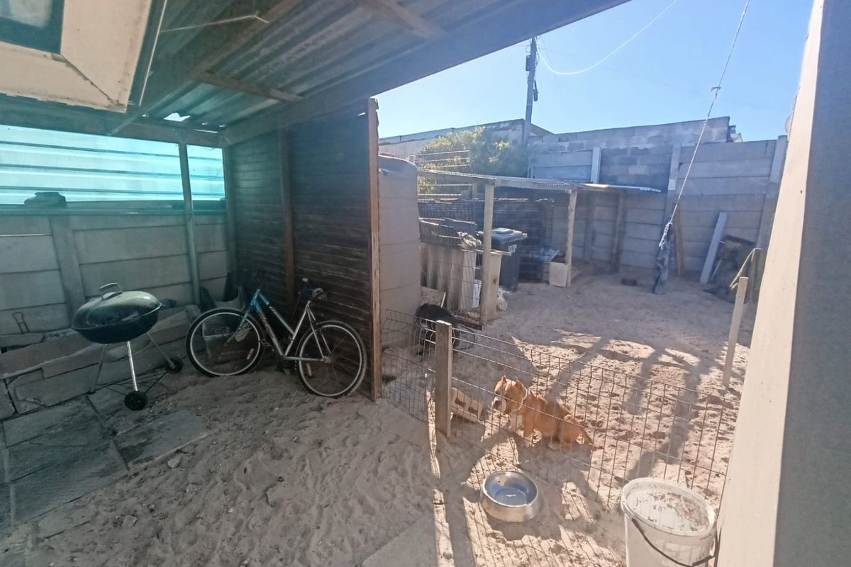 A rear outdoor area featuring a covered patio structure, concrete block boundary walls, and a water storage tank, currently with an unpaved sandy surface.