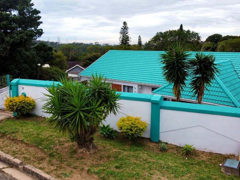 Front view of the property featuring a distinctive teal tiled roof, secure boundary wall, and gated entrance with landscaped garden area.