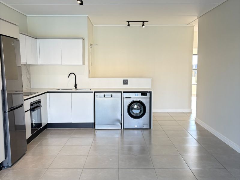 Modern open-plan kitchen featuring sleek white cabinetry, integrated appliances, tiled flooring, and contemporary track lighting in a clean, minimalist space.