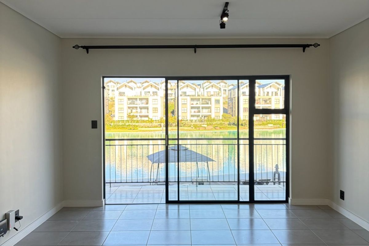 A bright, empty living room featuring neutral tiled flooring and large sliding glass doors that open onto a balcony with scenic water views.