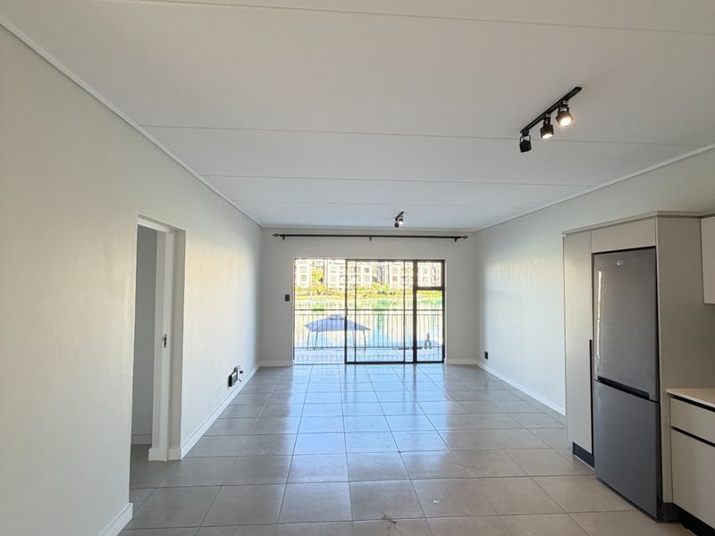 Spacious open-plan living and kitchen area featuring neutral tiled flooring, modern track lighting, and large sliding doors leading to a balcony with water views.