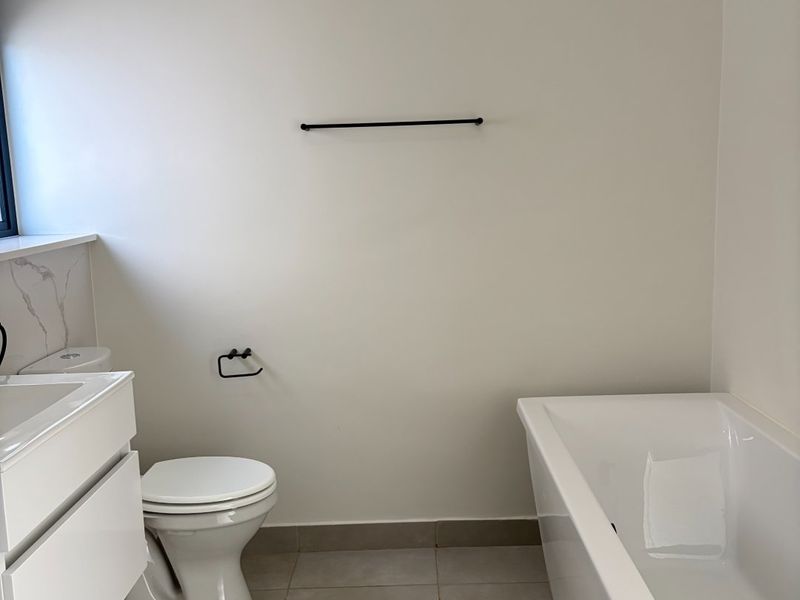 A clean, modern bathroom featuring a white vanity cabinet, integrated sink, toilet, and a bathtub with neutral floor tiling.