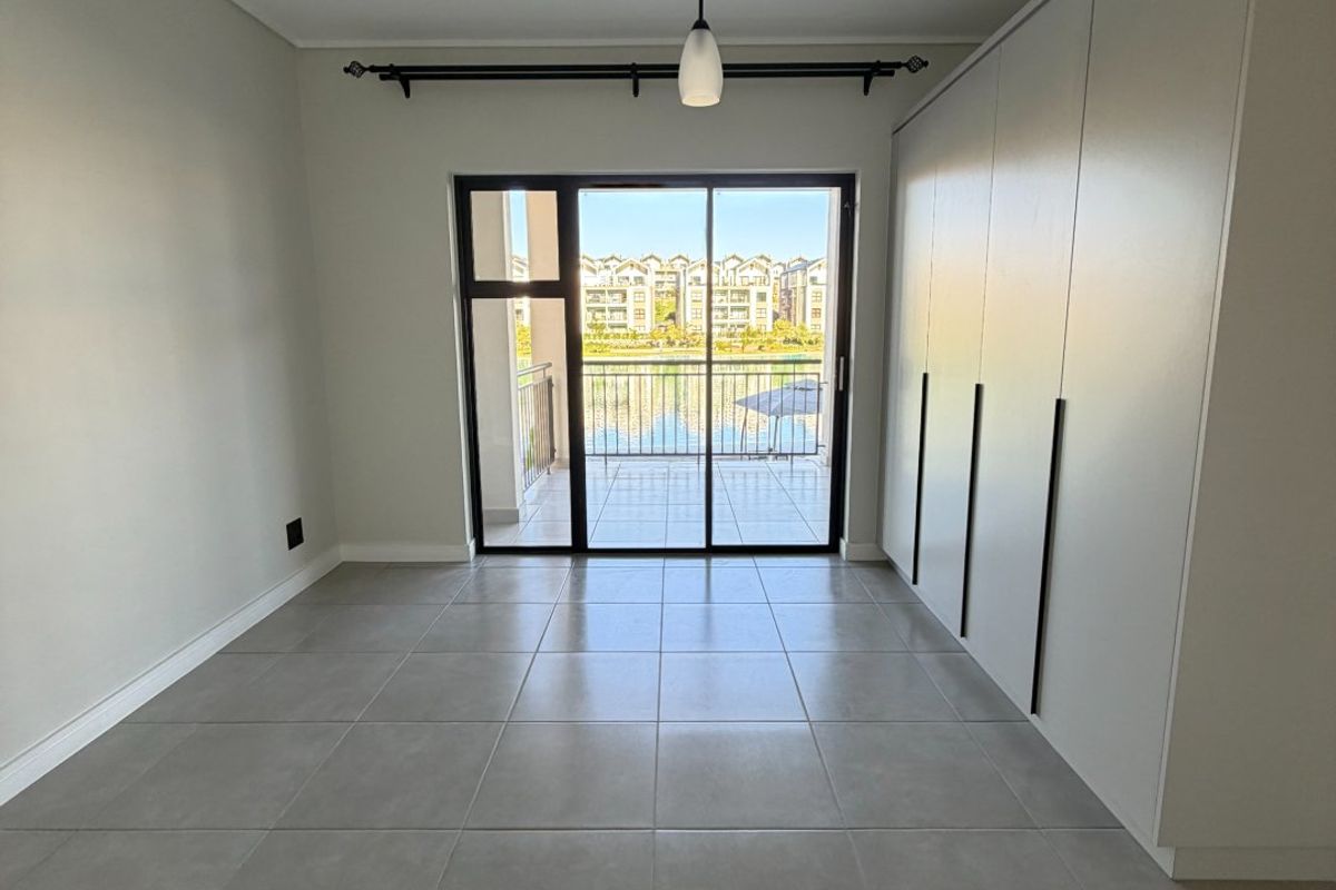A bright, empty bedroom featuring modern tiled flooring, built-in cupboards, and a sliding glass door leading to a balcony with scenic views.