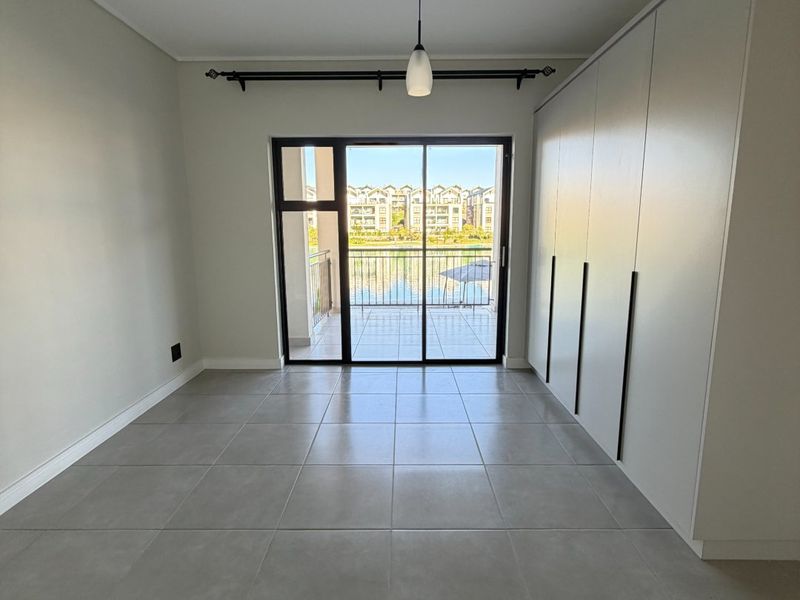 A bright, empty bedroom featuring modern tiled flooring, built-in cupboards, and a sliding glass door leading to a balcony with scenic views.