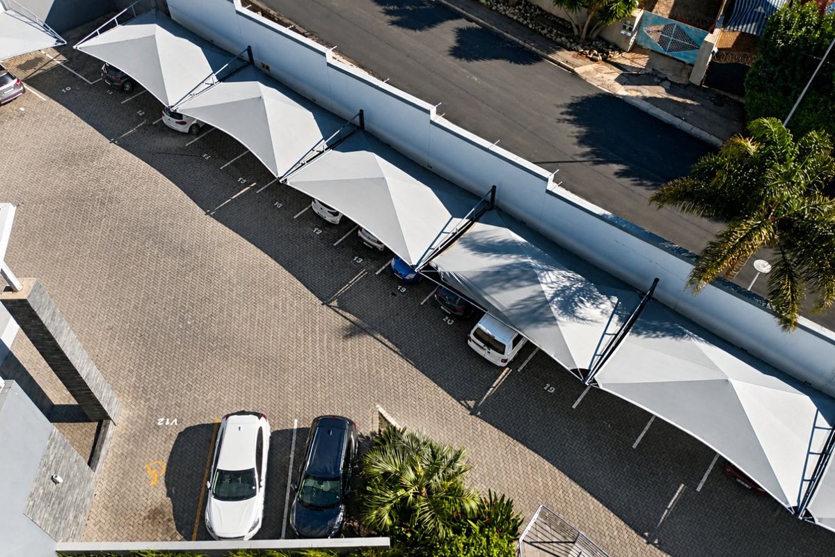 Aerial view of a paved apartment complex parking area featuring multiple shade cloth carports and a secure boundary wall.