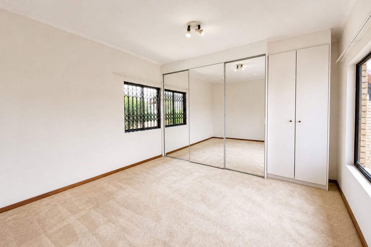 A bright, empty bedroom featuring neutral carpet flooring, built-in mirrored wardrobes, additional storage cupboards, and security gates on the windows.