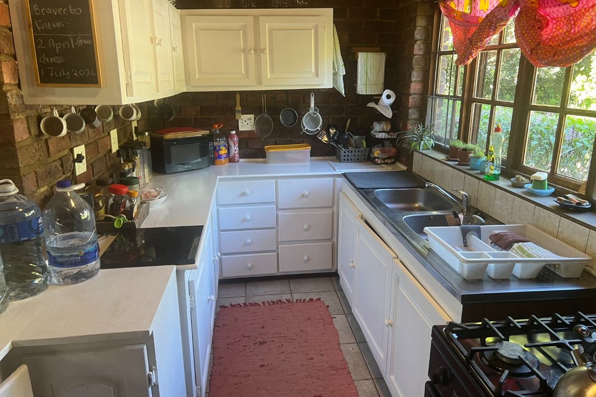 A compact kitchen featuring exposed brick walls, white cabinetry, a double bowl sink, and a built-in hob, offering a rustic aesthetic with potential for modernization.