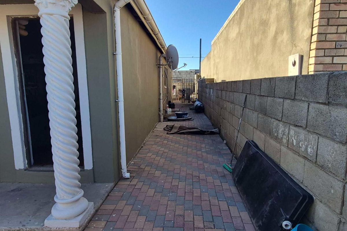 A narrow paved side passage featuring a decorative column, concrete block boundary wall, and exterior drainage infrastructure.
