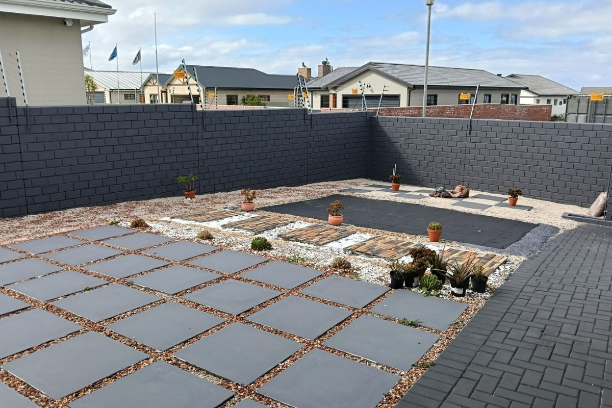 A low-maintenance rear courtyard featuring dark grey perimeter walls with electric fencing, stone landscaping, and modern square paving tiles.
