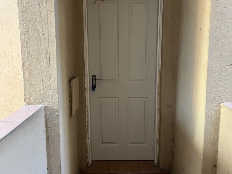 An exterior entrance area featuring tiled flooring and a panel door, showing visible signs of wear and plaster cracking above the door frame.