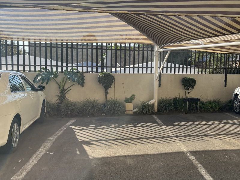 Covered parking bay featuring a striped shade cloth structure, asphalt surface, and secure perimeter fencing within the apartment complex.