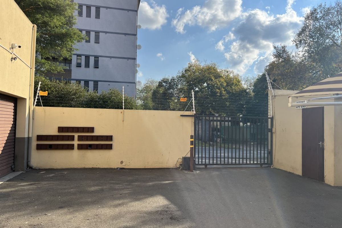 Exterior view of the apartment complex entrance featuring a secure gated access point, perimeter wall with electric fencing, and communal post boxes.