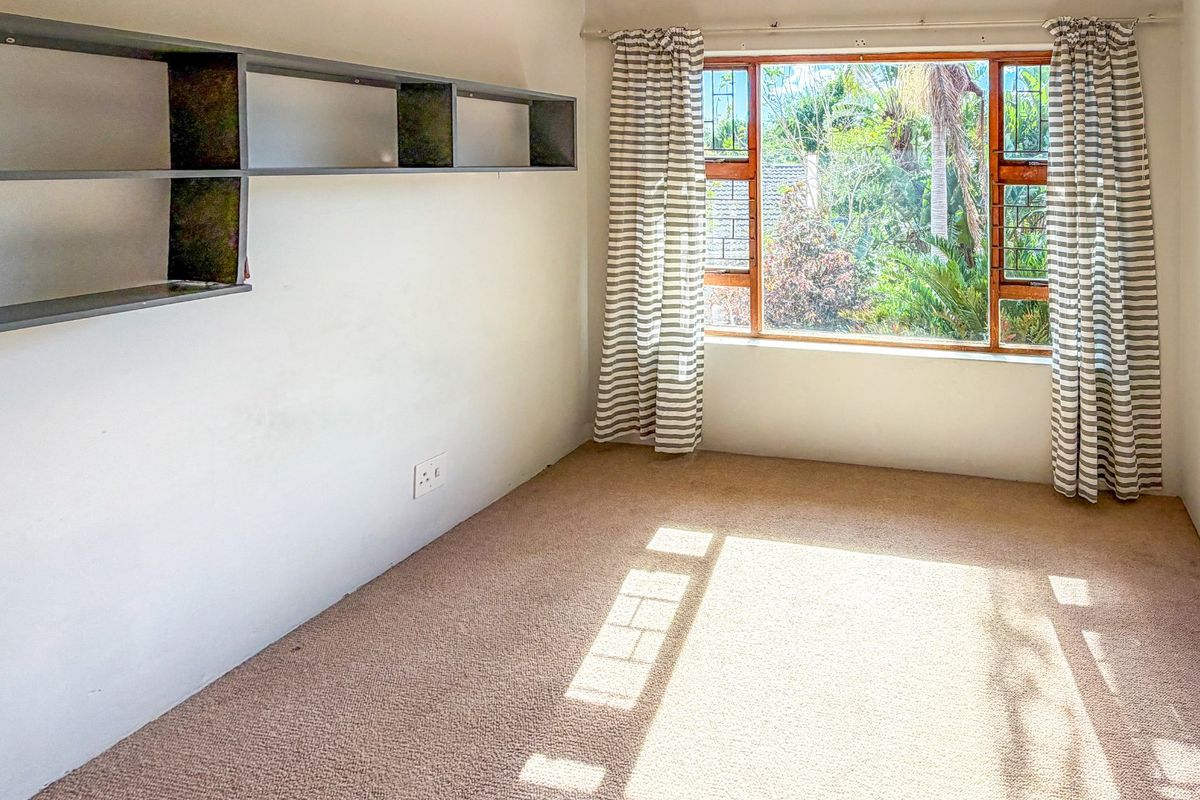 A bright, empty bedroom featuring built-in wall shelving, carpeted flooring, and a large window overlooking the garden.