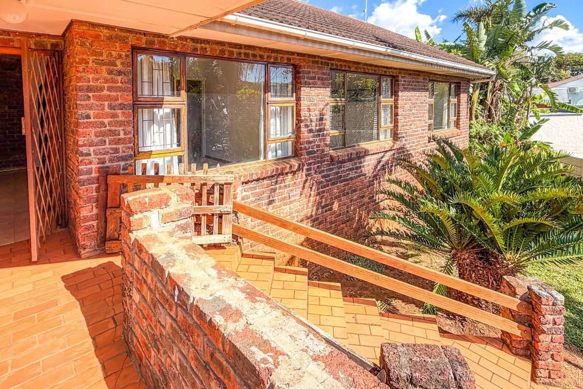 A rear exterior view featuring a face brick facade, a tiled patio area with a brick staircase, and timber handrails leading to the garden.