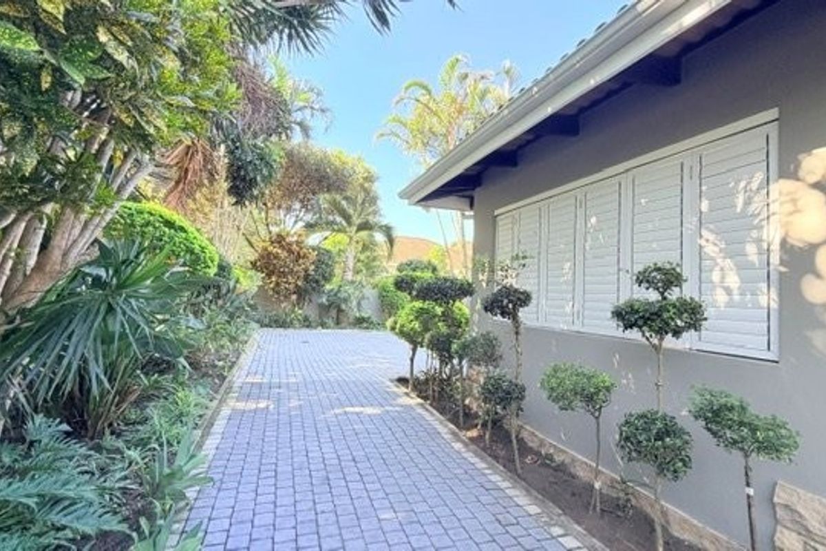 A well-maintained rear exterior view featuring a paved walkway, landscaped garden, and white shutters on the side of the townhouse.