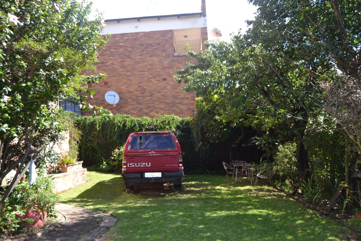 A rear garden area featuring a lawn, mature trees, and a brick building facade with a satellite dish.