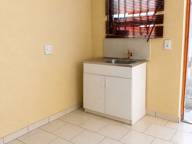 A functional laundry area featuring tiled flooring, a built-in vanity cabinet with a stainless steel sink, and a window with venetian blinds.