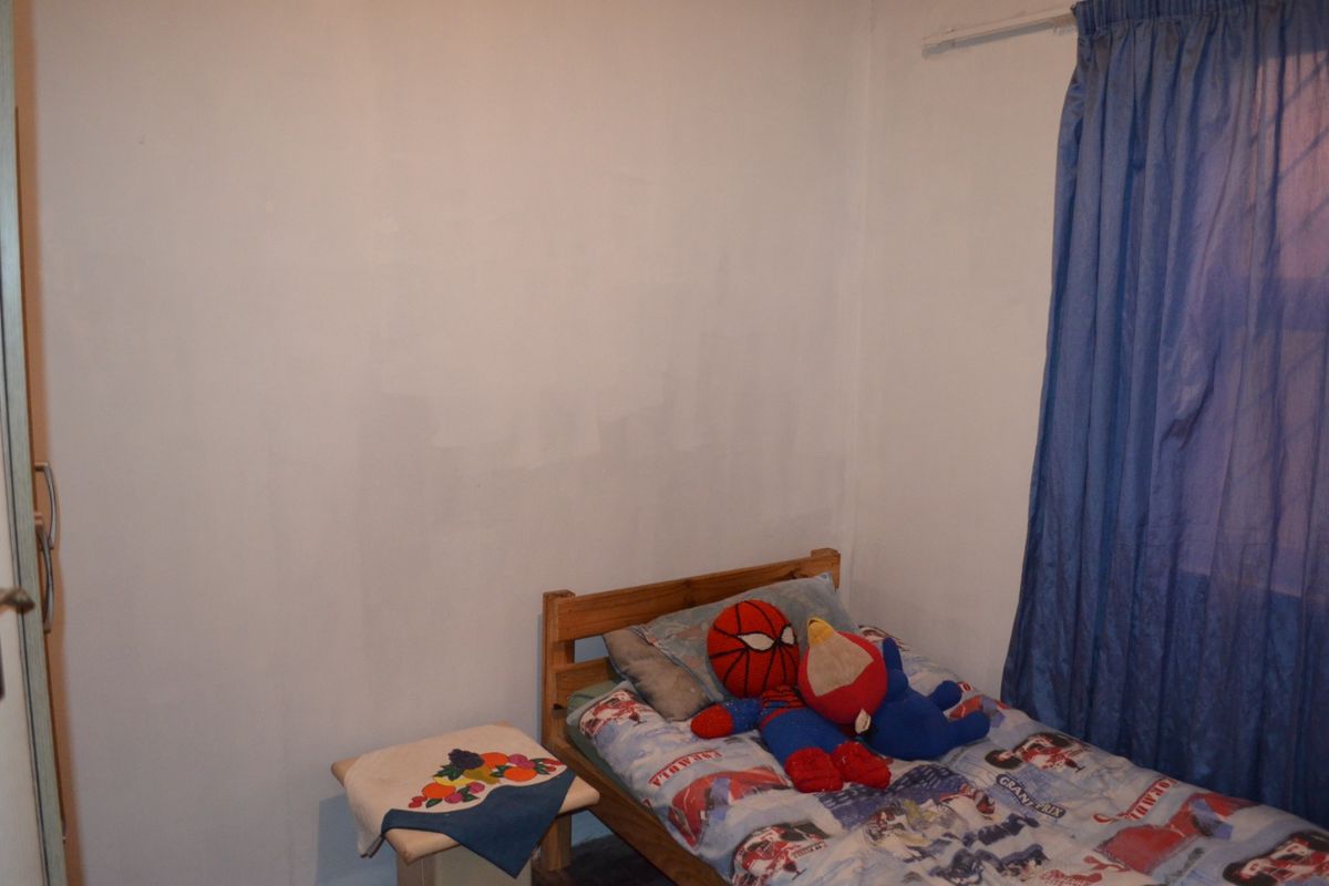 A compact bedroom featuring a built-in cupboard and a window with blue curtains. The walls show signs of uneven paint application.
