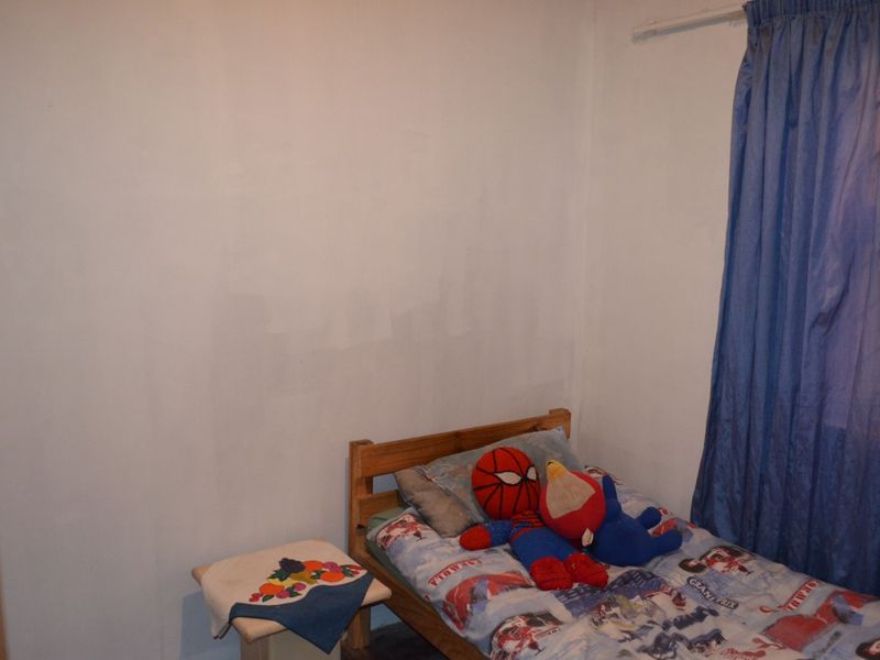 A compact bedroom featuring a built-in cupboard and a window with blue curtains. The walls show signs of uneven paint application.