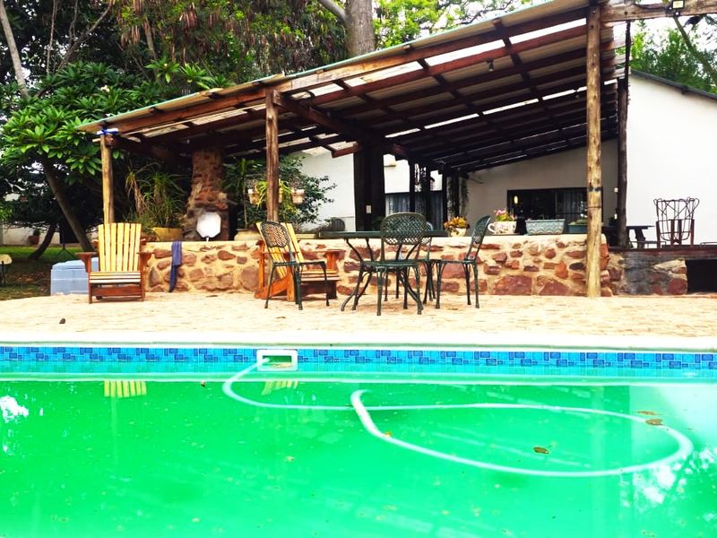Outdoor pool area featuring a paved deck, stone-clad patio wall, and a timber-framed covered entertainment space set within a garden environment.