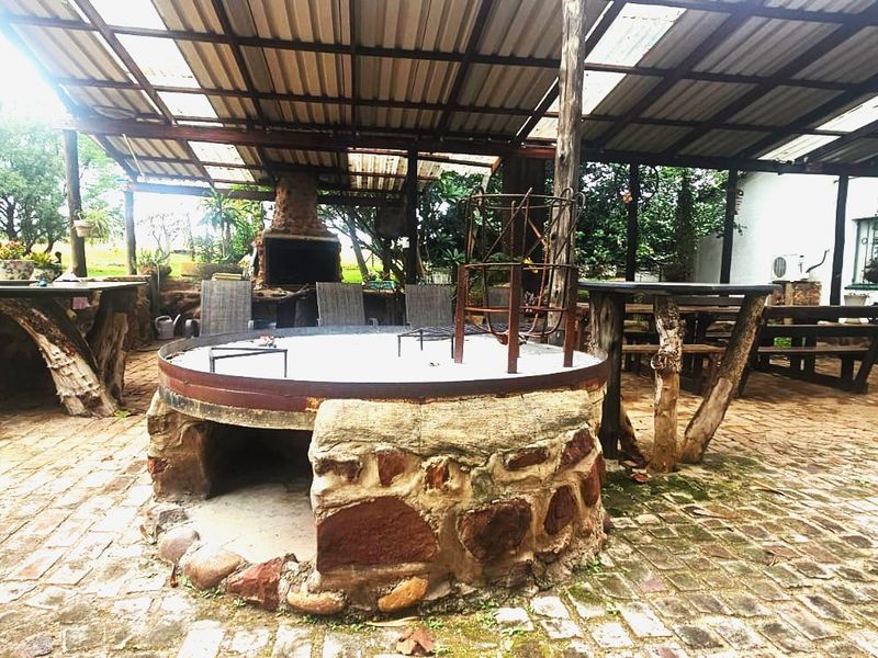 A rustic covered outdoor entertainment area featuring a built-in braai, a central stone fire pit, and brick paving under a corrugated roof structure.