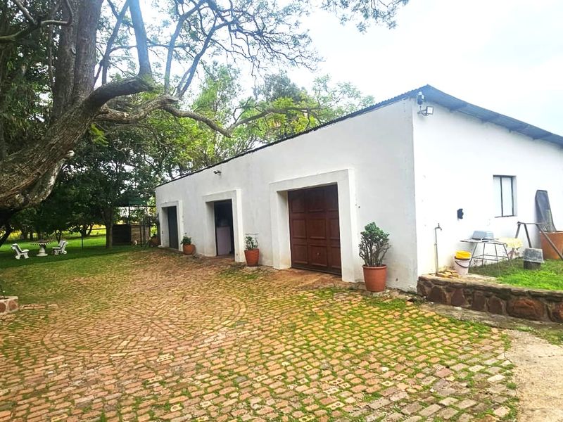 Rear view of a farm building featuring a cobblestone driveway, stone retaining wall, and timber garage door set within a mature garden.