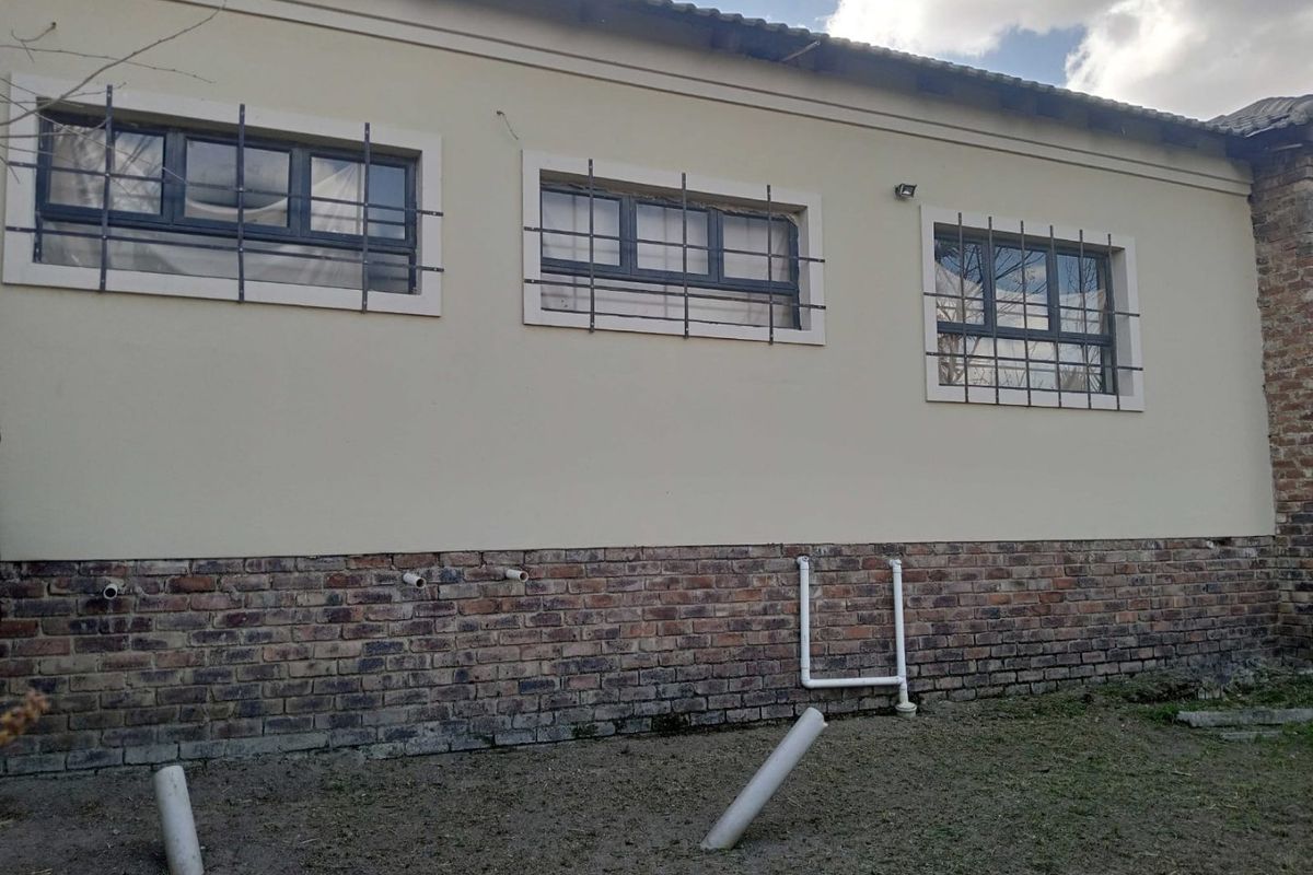 Rear exterior view of the property featuring a brick base, security-barred windows, and exposed plumbing infrastructure.