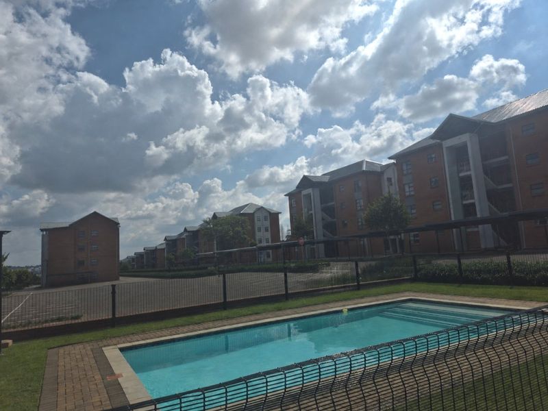 Communal swimming pool area featuring paved surrounds and metal fencing, set within a complex of multi-story brick apartment buildings.